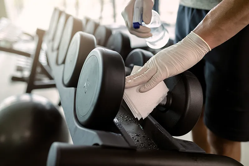 Nahaufnahme eines sportlichen Mannes, der in einem Fitnessstudio Hanteln mit Desinfektionsmittel reinigt.
